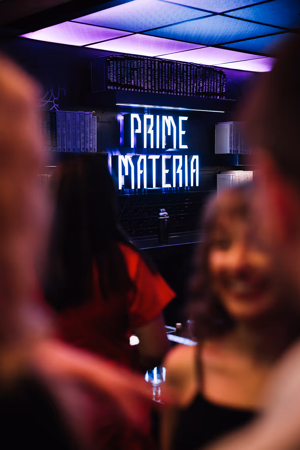 Neon sign reading 'PRIME MATERIA' glowing blue and purple with blurred people in foreground.