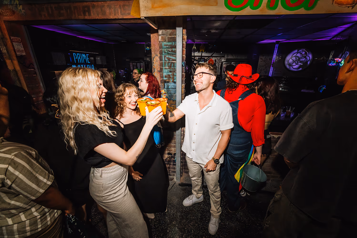 Group of people at a party, two smiling in foreground clinking colorful drinks together.
