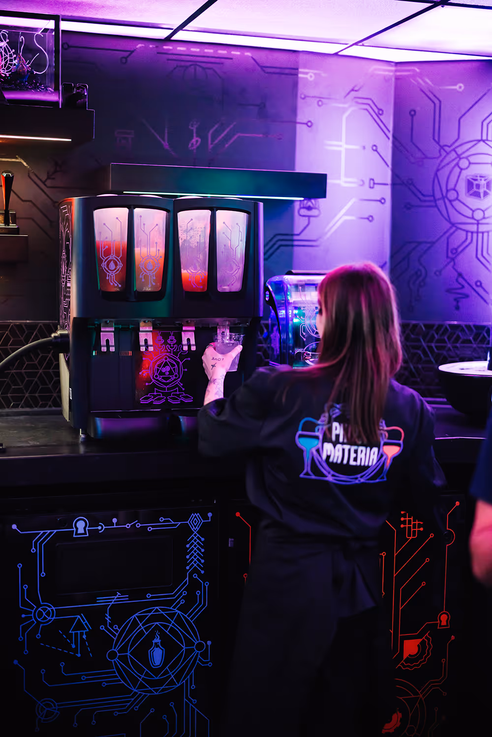 Person wearing a black apron fills a drink from a dispenser in a neon-lit tech-themed bar.