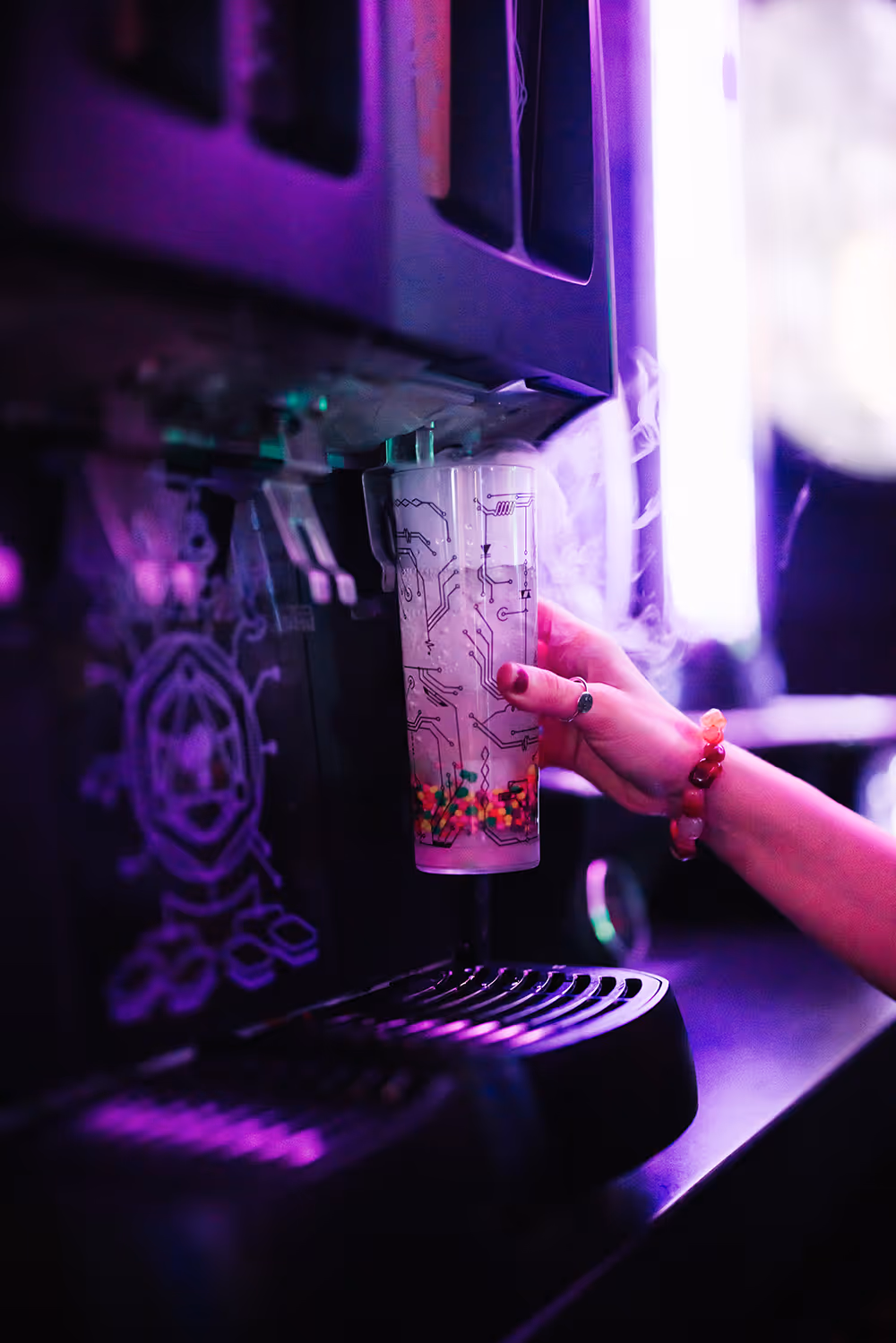 Hand holding a cup with circuit patterns and colorful balls under a purple-lit beverage dispenser. Prime Materia
