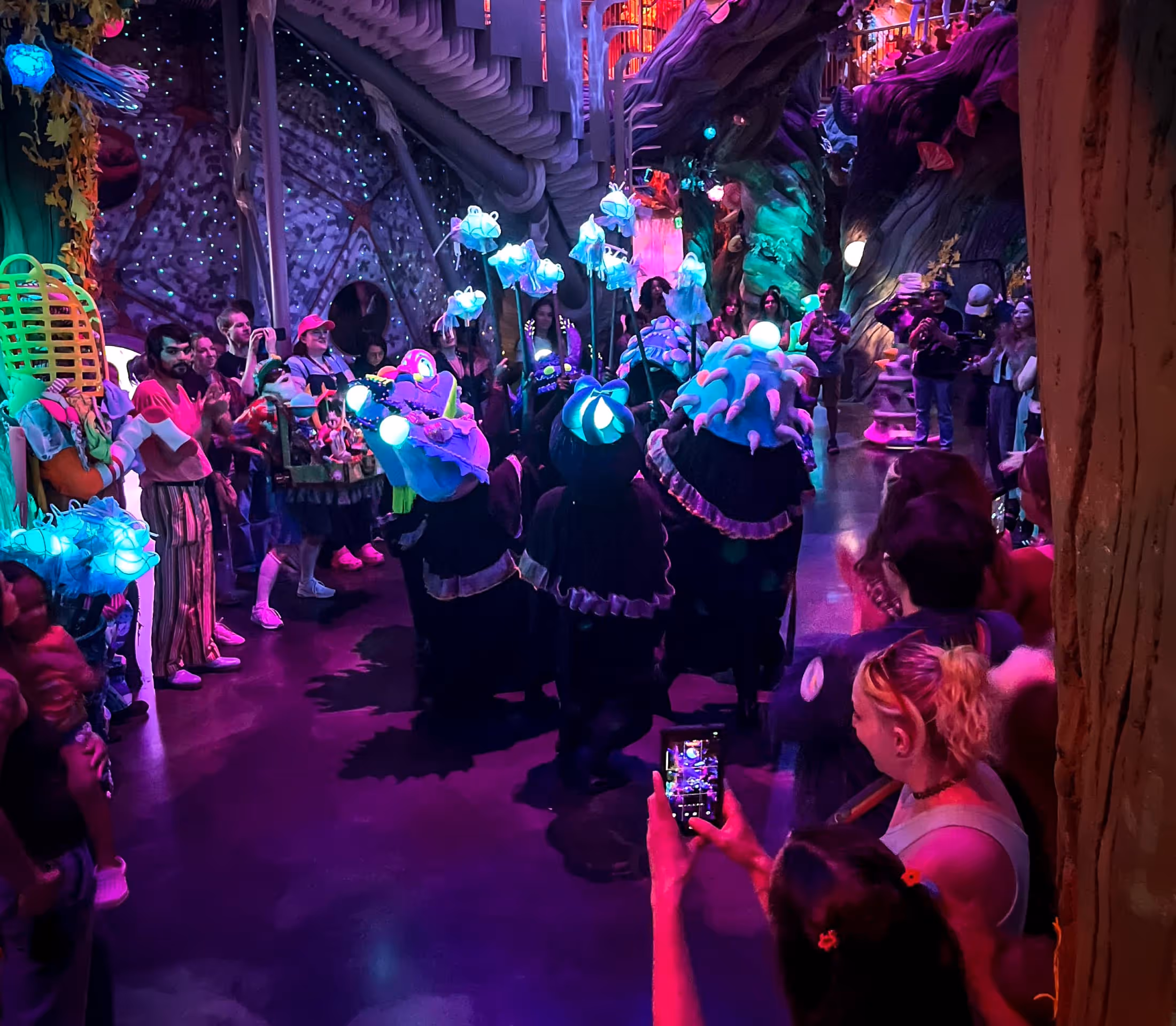 Crowd watches performers in glowing, colorful costumes at an indoor, fantasy-themed event.