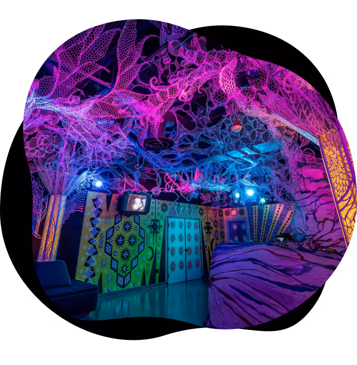 Room with neon-lit, web-like ceiling art and colorful patterned walls under blue and pink lights.